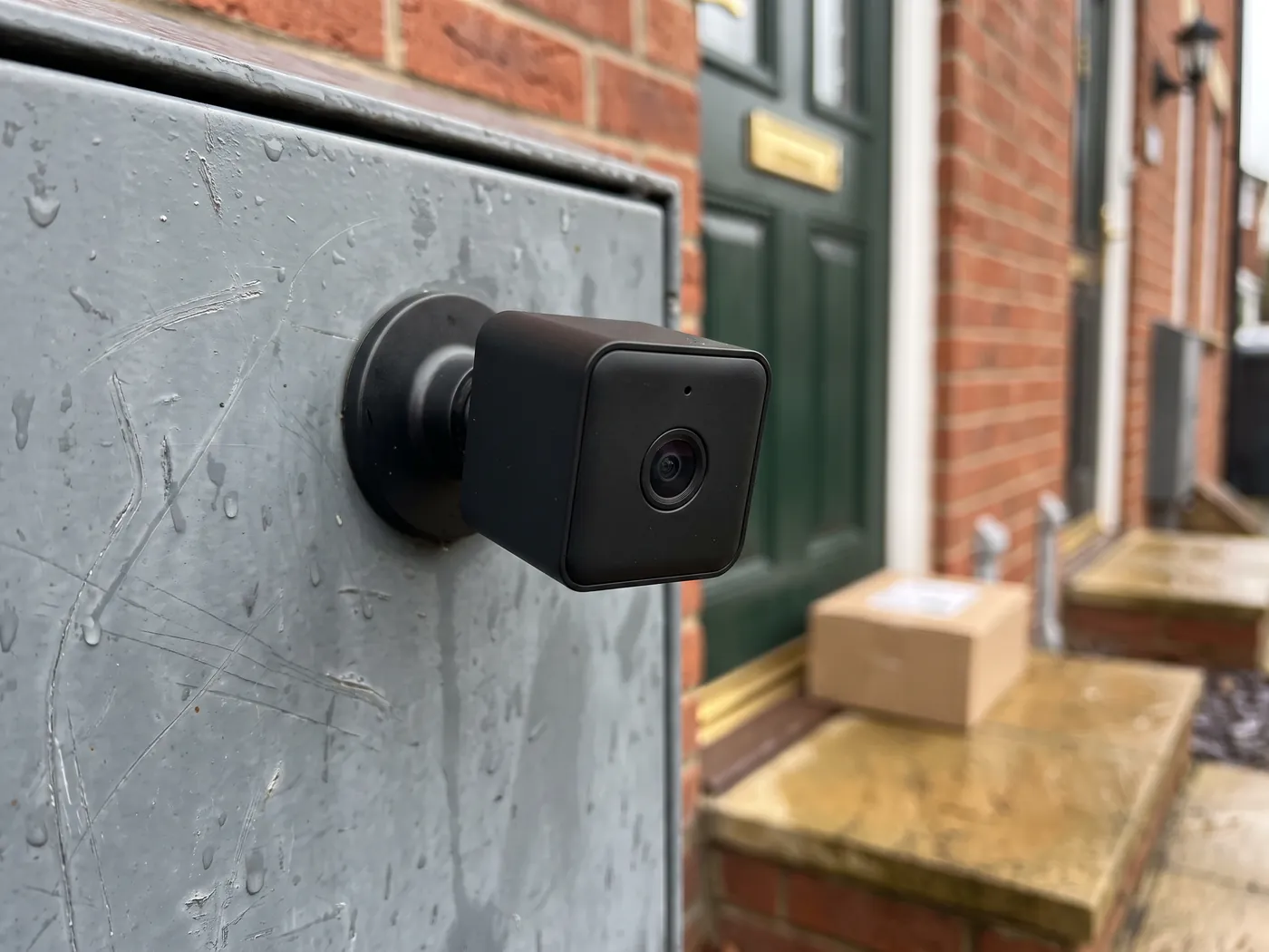 A small black mini camera on a magnetic base attached to a metal surface, with a parcel by the front door in the background